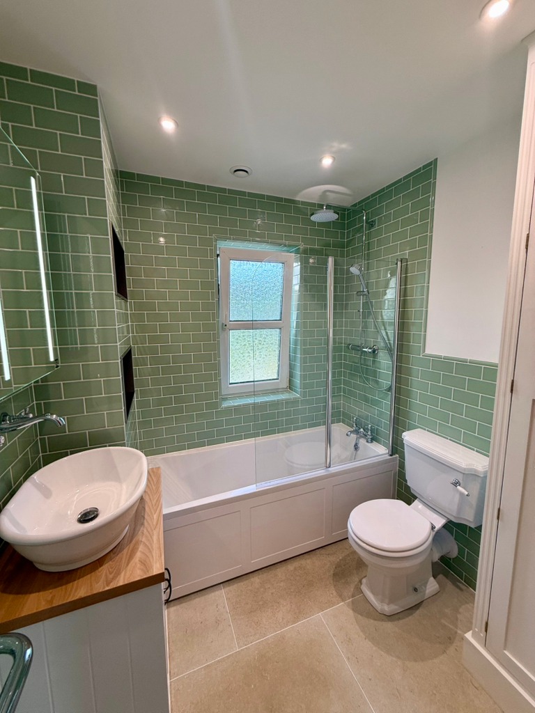 Green crackle-effect tiled bathroom with a bath below the window, brushed-brass fittings, countertop basin and toilet.